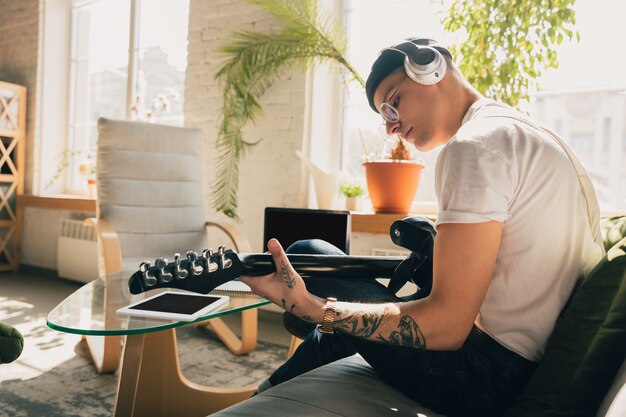 Hombre estudiando en casa durante cursos en línea o información gratuita por sí mismo. Se convierte en músico, guitarrista en aislamiento, cuarentena contra la propagación del coronavirus. Usando computadora portátil, teléfono inteligente, auriculares.