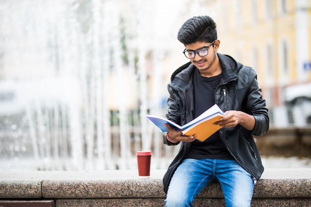 Hombre estudiante indio sosteniendo una pila de libros sentado cerca de la fuente en la calle