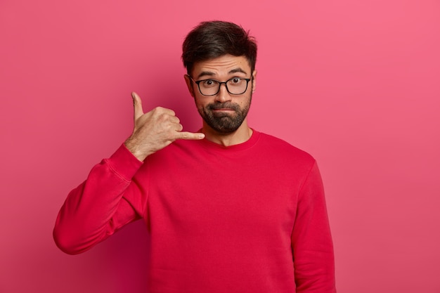 Un hombre europeo sin afeitar de aspecto serio hace un gesto de devolver la llamada, se mantiene siempre en contacto, usa gafas transparentes y un suéter rojo, pide un número de teléfono, aislado en una pared rosa.