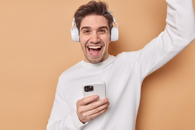 Un hombre europeo alegre y apuesto baila con triunfo escucha música a través de auriculares y se divierte usa tecnologías modernas para el entretenimiento usa un puente blanco casual aislado sobre fondo marrón.