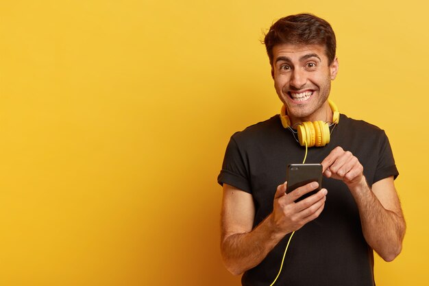 El hombre europeo alegre señala la pantalla del teléfono inteligente, usa auriculares amarillos, camiseta negra informal