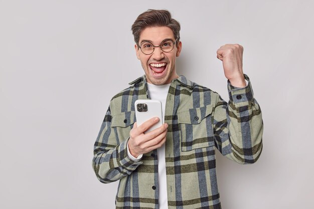 Un hombre europeo apuesto y alegre se siente contento de celebrar el juego en línea ganador, aprieta el puño con el triunfo, sostiene el teléfono móvil, usa gafas redondas y camisa a cuadros aislada sobre fondo blanco.