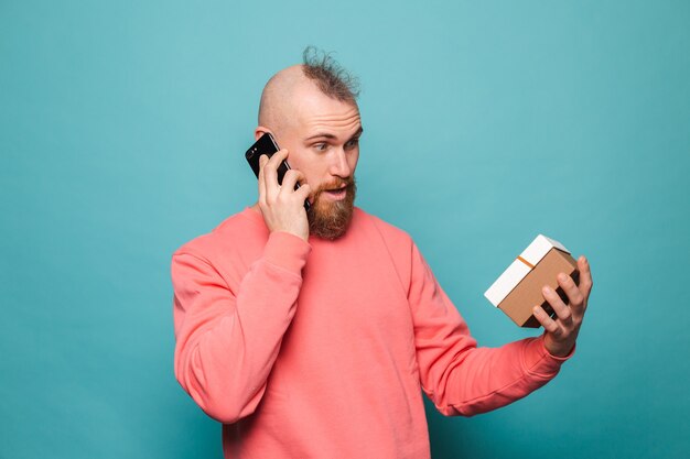 Hombre europeo barbudo en melocotón casual aislado, sosteniendo la caja de regalo hablar por teléfono móvil con cara de asombro sorprendido