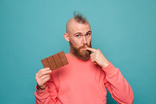 Hombre europeo barbudo en melocotón casual aislado, sosteniendo deliciosos dedos lamiendo chocolate