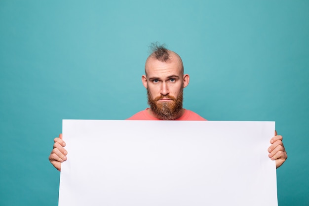 Hombre europeo barbudo en melocotón casual aislado, sosteniendo un tablero de papel vacío blanco con cara triste e infeliz