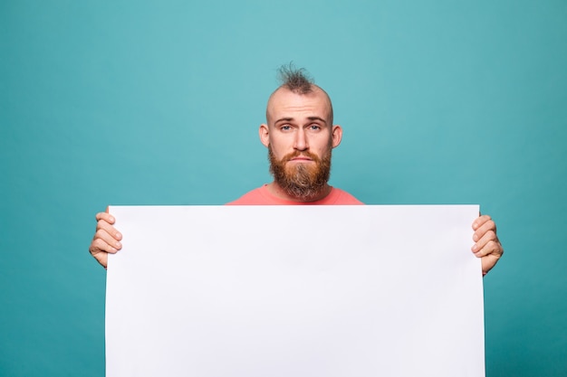 Hombre europeo barbudo en melocotón casual aislado, sosteniendo un tablero de papel vacío blanco con cara triste e infeliz