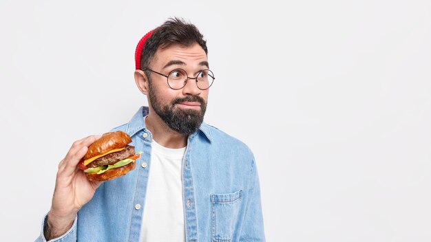 Hombre europeo barbudo sorprendido concentrado lejos sostiene hamburguesa come comida chatarra lleva gafas redondas y camisa de mezclilla
