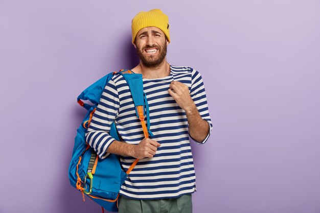 Hombre europeo disgustado con expresión molesta, aprieta los dientes nerviosamente, lleva mochila azul