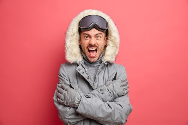 El hombre europeo emocional tiembla de frío y grita enojado mantiene los brazos cruzados vestido con chaqueta de invierno y va a hacer snowboard durante el día helado.