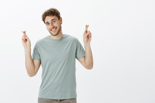 Hombre europeo encantador despreocupado en aretes con cabello rubio, inclinando la cabeza y sonriendo con expresión amistosa, levantando los dedos cruzados