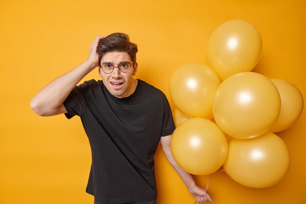 El hombre europeo indignado y disgustado mantiene la mano en la cabeza reacciona ante noticias desagradables usa una camiseta negra casual sostiene un montón de globos inflados aislados sobre fondo amarillo se prepara para la fiesta.