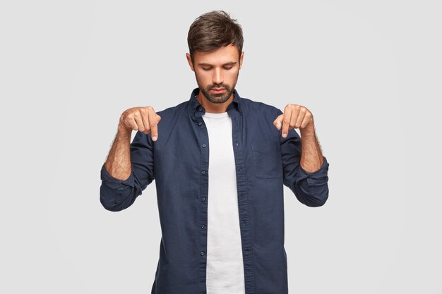 Hombre europeo joven serio concentrado con apariencia atractiva enfocada hacia abajo, indica con ambos dedos índices, tiene barba y bigote, viste camisa elegante, aislado sobre una pared blanca