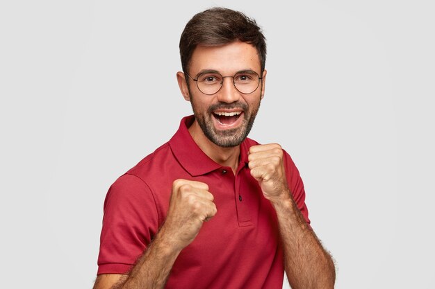 Hombre europeo positivo levanta los puños cerrados, listo para dar un puñetazo o pelear, sonríe ampliamente, vestido con una camiseta roja informal, se defiende aislado sobre una pared blanca. Gestos de hombre alegre enérgico