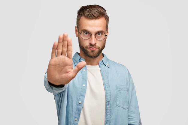 Un hombre europeo serio muestra un gesto de parada, exige algo, tiene una expresión facial estricta, usa gafas redondas y camisa formal, aislado sobre una pared blanca. Concepto de personas y lenguaje corporal