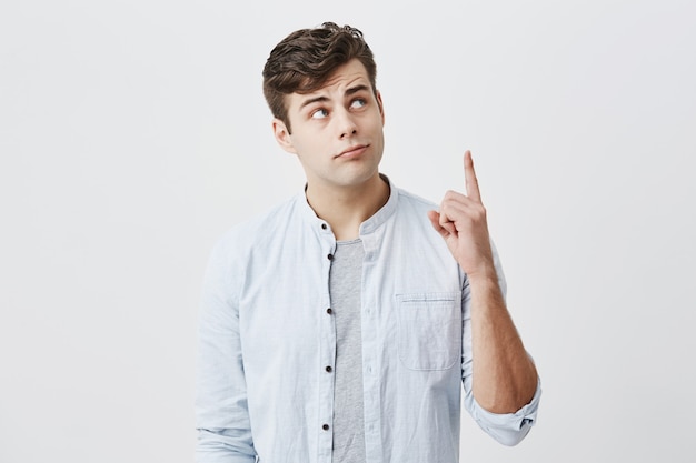 Hombre europeo serio y reflexivo con camisa azul claro sobre camiseta gris mirando hacia arriba, apuntando con el dedo índice hacia arriba con espacio para copiar su publicidad y contenido promocional. Concepto de publicidad.