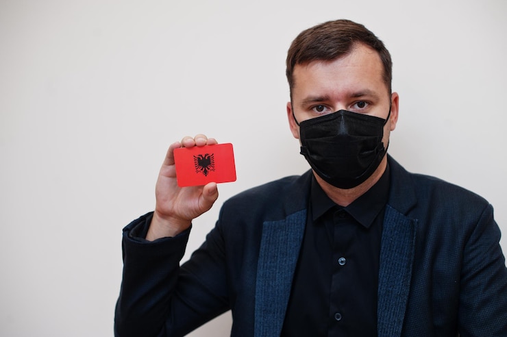 Foto gratuito el hombre europeo usa negro formal y protege la máscara facial sostiene la tarjeta de bandera de albania aislada sobre fondo blanco europa coronavirus covid concepto de país
