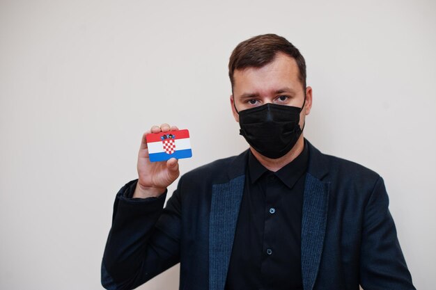 El hombre europeo usa negro formal y protege la máscara facial sostiene la tarjeta de bandera de Croacia aislada sobre fondo blanco Europa coronavirus Covid concepto de país