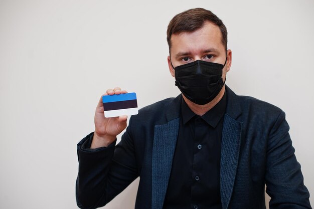 El hombre europeo usa negro formal y protege la máscara facial sostiene la tarjeta de bandera de Estonia aislada sobre fondo blanco Europa coronavirus Covid concepto de país