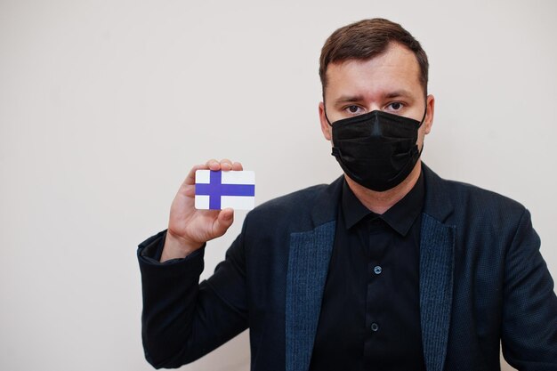 El hombre europeo usa negro formal y protege la máscara facial sostiene la tarjeta de bandera de Finlandia aislada sobre fondo blanco Europa coronavirus Covid concepto de país