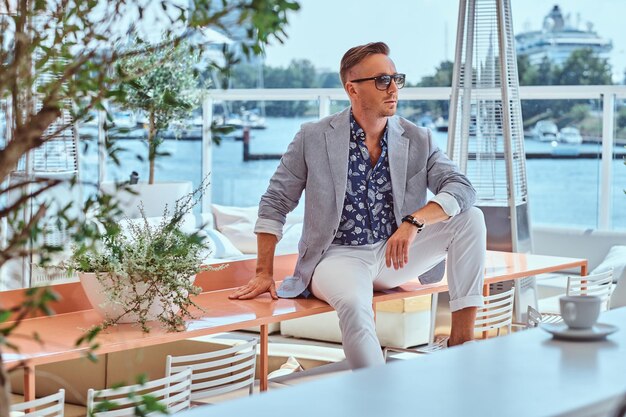 Un hombre exitoso y elegante vestido con ropa moderna y elegante sentado en una mesa en un café al aire libre con el telón de fondo del muelle de la ciudad.