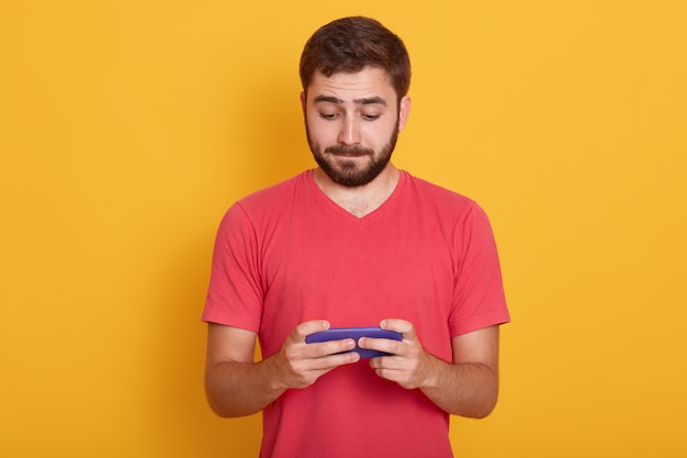 hombre con expresión facial tranquila, camiseta roja casual, jugando juegos en línea en el teléfono inteligente o revisando su red social, posando aislado en amarillo. Concepto de tecnología