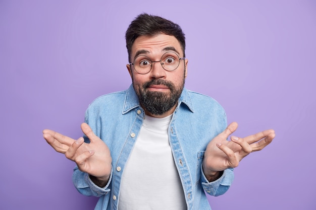 El hombre extiende las palmas de las manos se siente despistado e incierto, no puede elegir, usa anteojos redondos, camisa vaquera, poses