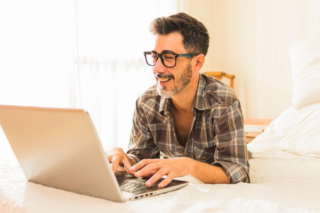 Hombre feliz acostado en una cama acogedora usando la laptop