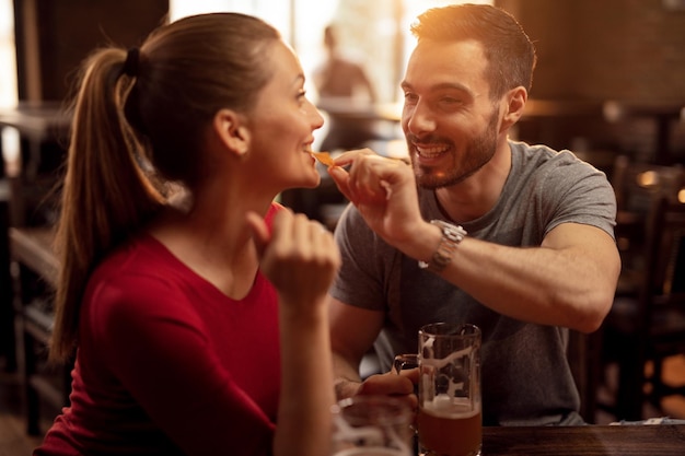 Un hombre feliz alimentando a su novia con chips de nacho mientras beben cerveza juntos en un pub