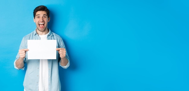 Foto gratuita un hombre feliz y atractivo que muestra un trozo de papel para su logotipo de pie asombrado contra el fondo azul