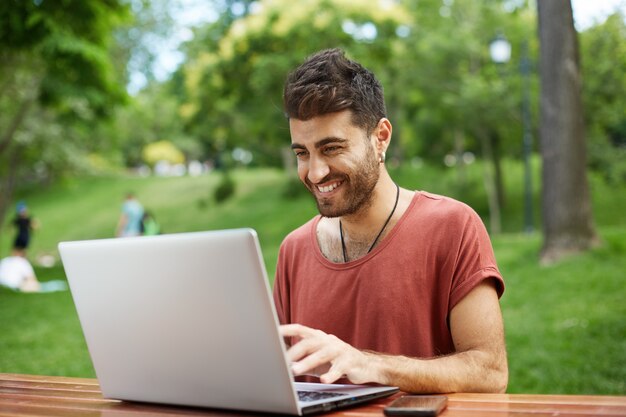 Hombre feliz atractivo que usa la computadora portátil en el parque, viendo videos o podcast al aire libre