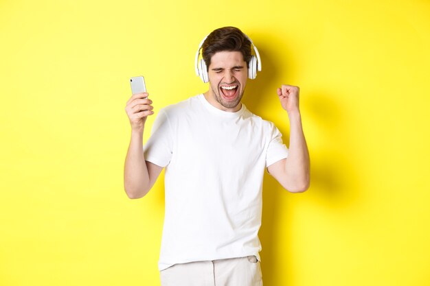 Hombre feliz bailando y escuchando música en auriculares, sosteniendo el teléfono celular móvil, de pie contra el fondo amarillo.