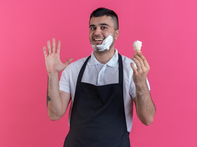 Hombre feliz barbero en delantal poniendo espuma de afeitar en su rostro con brocha de afeitar mirando confiado de pie sobre fondo rosa