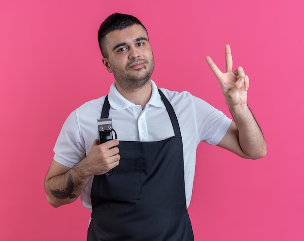 Hombre feliz barbero en delantal sosteniendo recortadora mirando a la cámara sonriendo alegremente mostrando v-sign de pie sobre fondo rosa