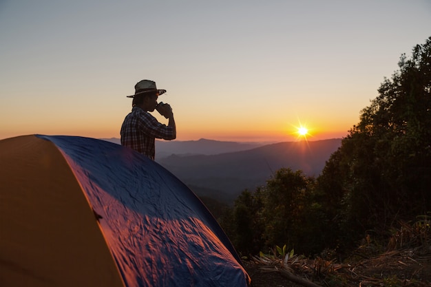 Hombre feliz con bebida, cerca de la tienda alrededor de las montañas bajo la luz del atardecer