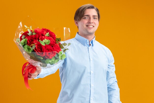 Hombre feliz en blueshirt sosteniendo un ramo de rosas rojas mirando a la cámara sonriendo alegremente el día de San Valentín concepto de pie sobre la pared naranja