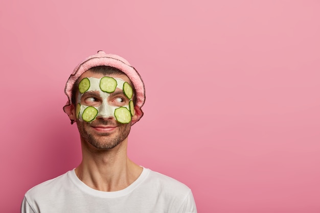 El hombre feliz de buen carácter usa mascarilla y pepinos, disfruta del procedimiento de spa matutino, quiere lucir renovado