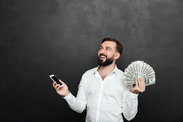 hombre feliz en camisa blanca ganando mucho dinero en efectivo usando su teléfono móvil y mirando hacia arriba en gris oscuro