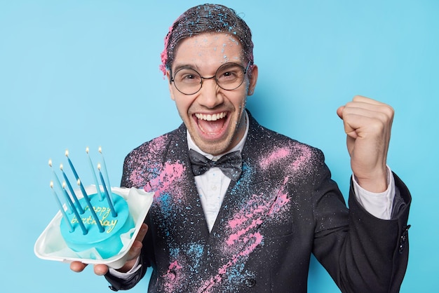 Hombre feliz celebra cumpleaños con amigos en arty sostiene pastel festivo aprieta el puño con triunfo viste traje formal expresa emociones positivas aisladas sobre fondo azul Gente y celebración