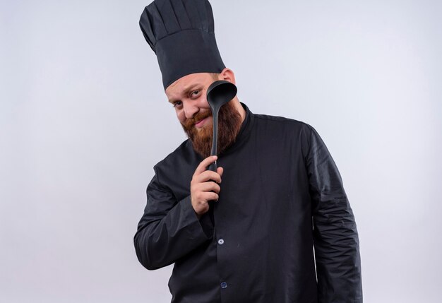 Un hombre feliz chef barbudo en uniforme negro mostrando cucharón negro mientras mira a la cámara en una pared blanca