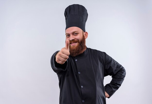 Un hombre feliz chef barbudo en uniforme negro mostrando los pulgares hacia arriba mientras mira a la cámara en una pared blanca
