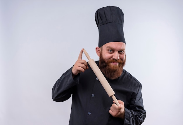 Un hombre feliz chef barbudo en uniforme negro que muestra el rodillo mientras mira a la cámara en una pared blanca