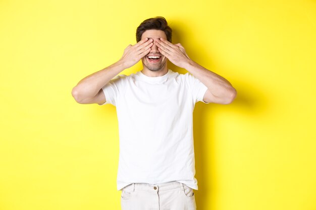 Hombre feliz cierra los ojos y espera sorpresa, sonriendo divertido, de pie sobre fondo amarillo.