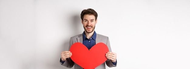 Foto gratuita hombre feliz confesándose enamorado mostrando corte de corazón y sonriendo a la cámara de pie en traje romántico