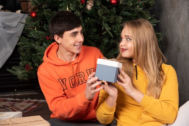 Hombre feliz dando caja de regalo de Navidad a su novia cerca del árbol de Navidad.