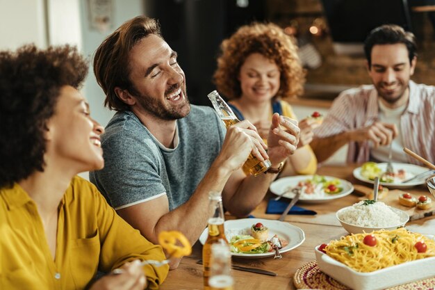 Hombre feliz divirtiéndose mientras almuerza y bebe cerveza con amigos en casa