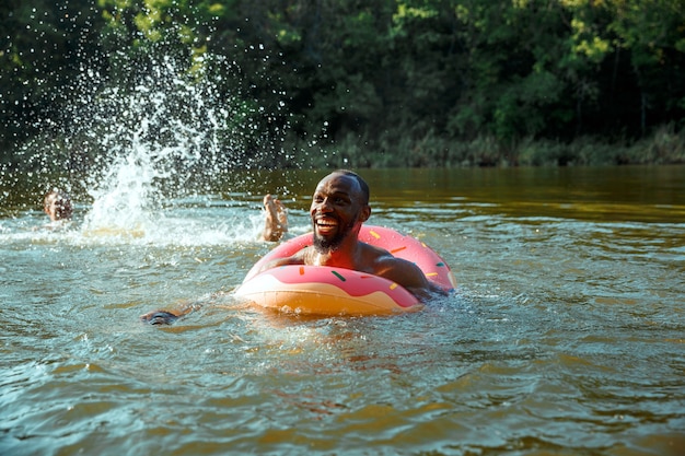 Hombre feliz divirtiéndose mientras se ríe y nada en el río. Modelos masculinos alegres con anillo de goma como un donut en la orilla del río en un día soleado. Verano, amistad, resort, concepto de fin de semana.