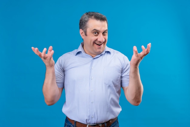 Hombre feliz y emocionado en camisa a rayas azul levantando las manos con expresión complacida en un espacio azul