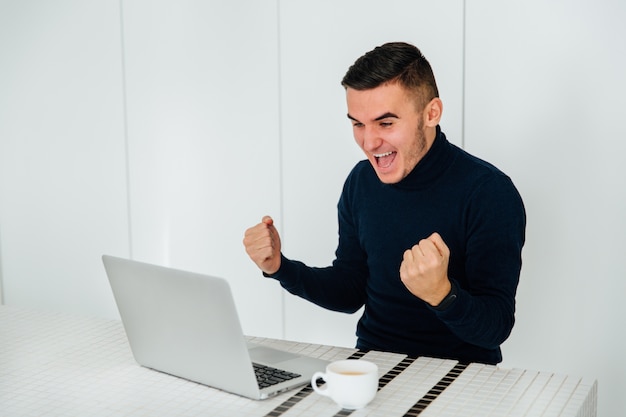 El hombre feliz emocionado que muestra los puños, celebra su éxito después de ganar en línea.