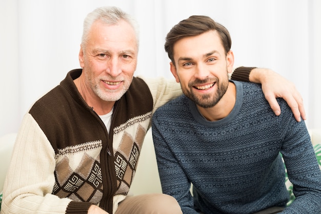 Hombre feliz envejecido que abraza al individuo sonriente joven en el sofá