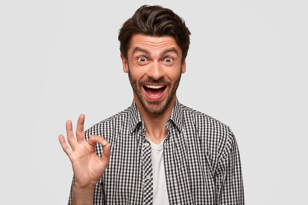 El hombre feliz feliz hace un buen gesto, muestra que todo está bien, dice que esté tranquilo, hace gestos en el interior contra la pared blanca